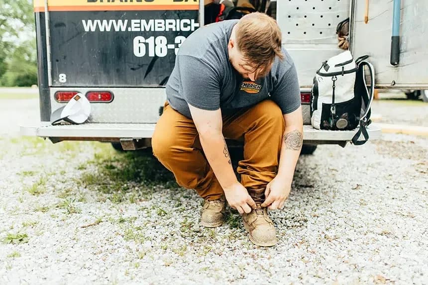 Embrich Plumbing, Heating & Air Conditioning service expert crouching to perform plumbing work at an outdoor job site in Collinsville