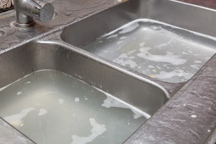 Clogged double-basin stainless steel kitchen sink with standing water and debris, requiring professional drain cleaning services from Embrich Plumbing