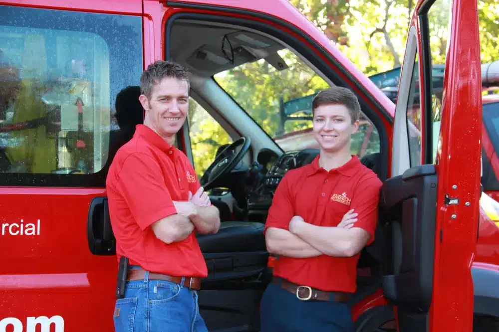 Two Embrich Plumbing, Heating & Air Conditioning technicians standing beside branded service van, smiling
