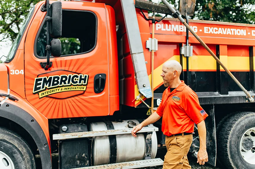 Embrich Plumbing, Heating & Air Conditioning service expert standing beside large orange branded work truck at job site in Collinsville